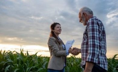 Senior man farmer shaking hands with young pretty woman with notebook in corn field. Insurance in agribusiness concept