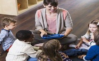 A high angle view of a woman teaching a multiracial group of preschoolers or kindergarten children, holding a digital tablet.  They are in a classroom, sitting in a circle on a wooden floor.  There are five girls and boys facing the teacher who is sitting cross-legged and smiling.